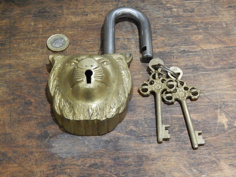 Puede incluir: Un candado de lat&oacute;n vintage con un dise&ntilde;o de cabeza de le&oacute;n y un grillete de acero abierto. Dos llaves ornamentadas est&aacute;n al lado del candado. El candado est&aacute; sobre una superficie de madera.