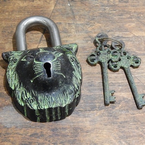Puede incluir: Un candado de estilo vintage con dise&ntilde;o de cabeza de le&oacute;n, grillete plateado y ojo de cerradura. El candado es negro con detalles verdes, acompa&ntilde;ado de dos llaves ornamentadas.