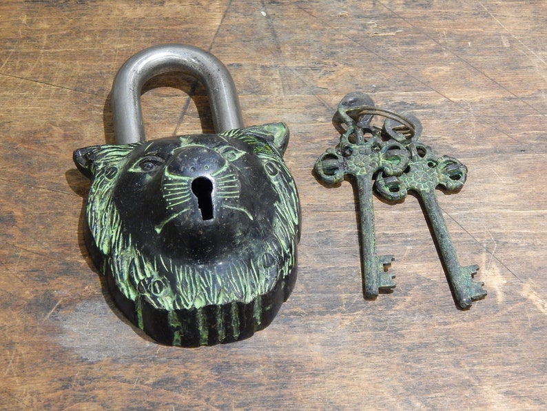 Puede incluir: Un candado de estilo vintage con dise&ntilde;o de cabeza de le&oacute;n, grillete plateado y ojo de cerradura. El candado es negro con detalles verdes, acompa&ntilde;ado de dos llaves ornamentadas.