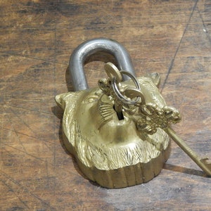 Puede incluir: Un candado de lat&oacute;n vintage con un dise&ntilde;o de cabeza de le&oacute;n, con un grillete plateado y un juego de llaves. Las llaves est&aacute;n insertadas en la cerradura. El candado est&aacute; sobre una superficie de madera.