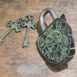 Puede incluir: Un candado de estilo antiguo con una cara decorativa y dos llaves a juego. El candado es negro con detalles verdes, y las llaves son de un verde a juego. El candado y las llaves est&aacute;n sobre una superficie de madera.