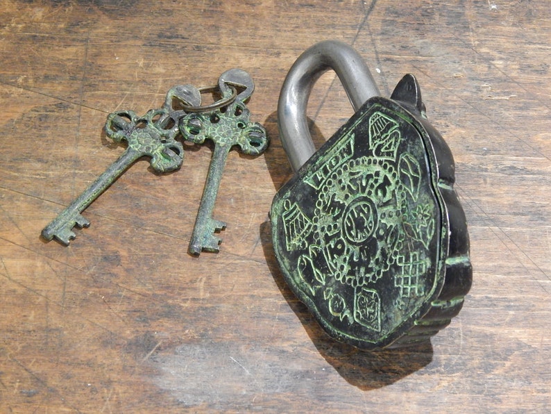 Puede incluir: Un candado de estilo antiguo con una cara decorativa y dos llaves a juego. El candado es negro con detalles verdes, y las llaves son de un verde a juego. El candado y las llaves est&aacute;n sobre una superficie de madera.