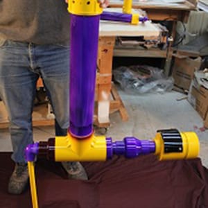 Könnte beinhalten: Ein violettes und gelbes PVC-Rohr-Gerät mit einem Griff und einer Düse. Das Gerät steht auf einer braunen Oberfläche.