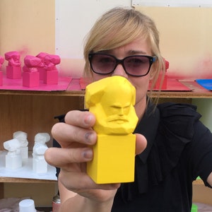 May include: A yellow Karl Marx bust is held up in front of a person wearing sunglasses. Other busts in pink, white, and blue are on shelves in the background. The busts are geometric and stylized.