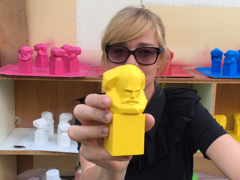 May include: A yellow Karl Marx bust is held up in front of a person wearing sunglasses. Other busts in pink, white, and blue are on shelves in the background. The busts are geometric and stylized.