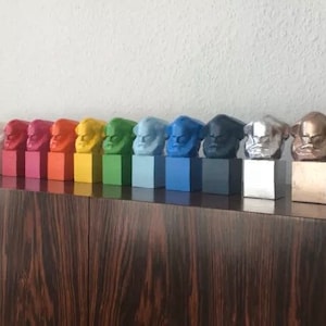 May include: A row of colourful bust sculptures, each mounted on a square base. The busts are painted in various colours, including black, white, red, orange, yellow, green, blue, and metallic silver and gold. The sculptures are displayed on a dark wood surface.
