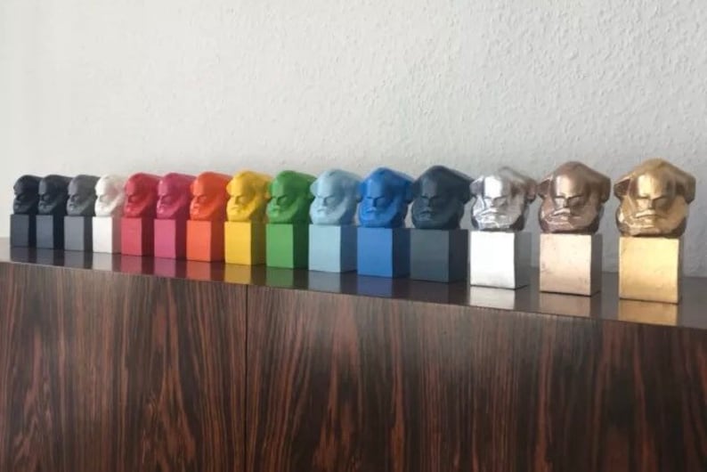 May include: A row of colourful bust sculptures, each mounted on a square base. The busts are painted in various colours, including black, white, red, orange, yellow, green, blue, and metallic silver and gold. The sculptures are displayed on a dark wood surface.