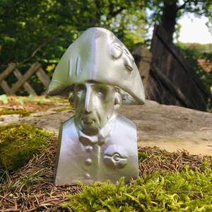 May include: A silver bust of a man wearing a tricorn hat. The bust is sitting on a bed of moss and pine needles.