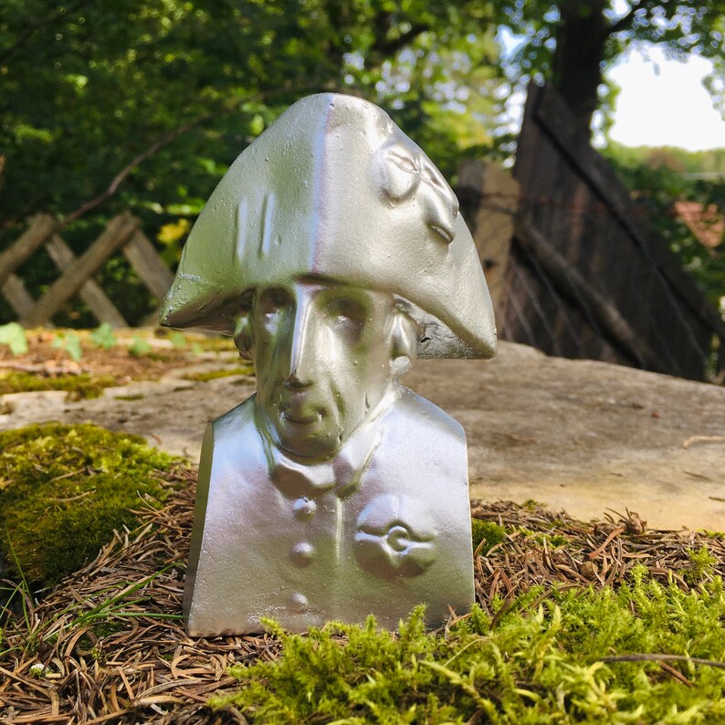 May include: A silver bust of a man wearing a tricorn hat. The bust is sitting on a bed of moss and pine needles.