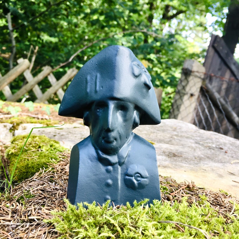 May include: A dark blue bust of a person wearing a bicorne hat. The bust is made of a smooth, matte material and has a detailed face. The bust is sitting on a bed of moss and pine needles.