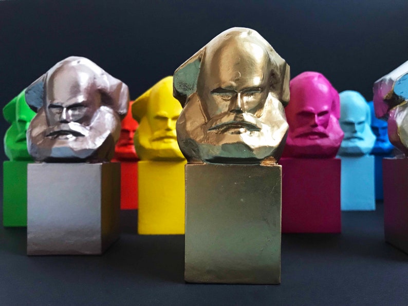 May include: A selection of colourful busts of a bearded man, each on a square base. The busts are painted in various colours, including gold, silver, yellow, pink, blue, and green. The background is dark.