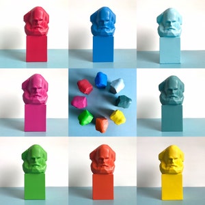 May include: A selection of brightly coloured Karl Marx bust sculptures on square plinths. The busts and plinths are painted in various colours, including red, blue, pink, green, and yellow. Additional colourful rock-shaped sculptures are also present.