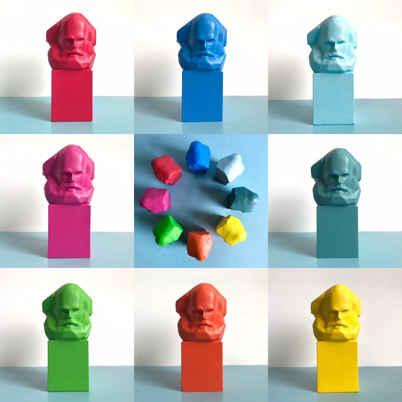 May include: A selection of brightly coloured Karl Marx bust sculptures on square plinths. The busts and plinths are painted in various colours, including red, blue, pink, green, and yellow. Additional colourful rock-shaped sculptures are also present.
