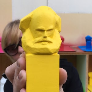 May include: A vibrant yellow sculpture of a bust with a beard and a serious expression. The sculpture is held in front of a person. Other similar sculptures in pink and blue are visible in the background.