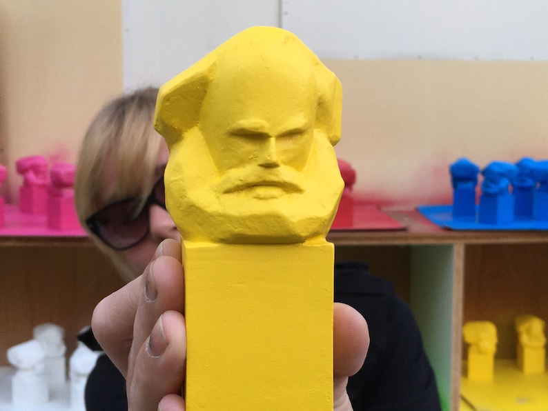 May include: A vibrant yellow sculpture of a bust with a beard and a serious expression. The sculpture is held in front of a person. Other similar sculptures in pink and blue are visible in the background.