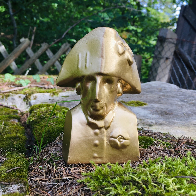 May include: A gold-painted bust of a person wearing a hat with a decorative design. The bust is sitting on a bed of moss and pine needles.