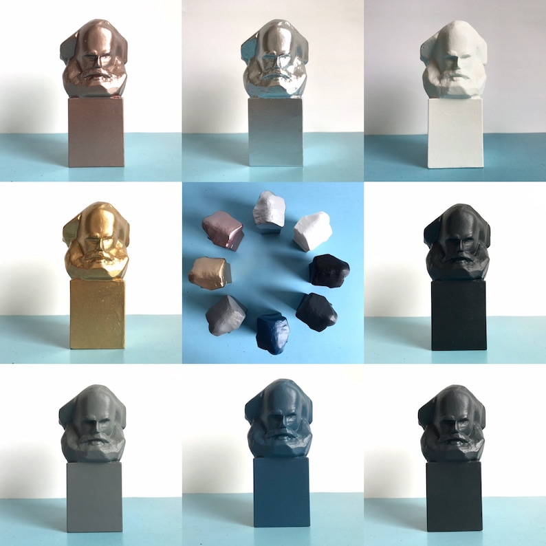 May include: A selection of Karl Marx bust sculptures in diverse colours, including metallic bronze, silver, and gold, alongside matte white, black, and grey. The busts are mounted on rectangular plinths, with additional smaller, irregularly shaped sculptures.