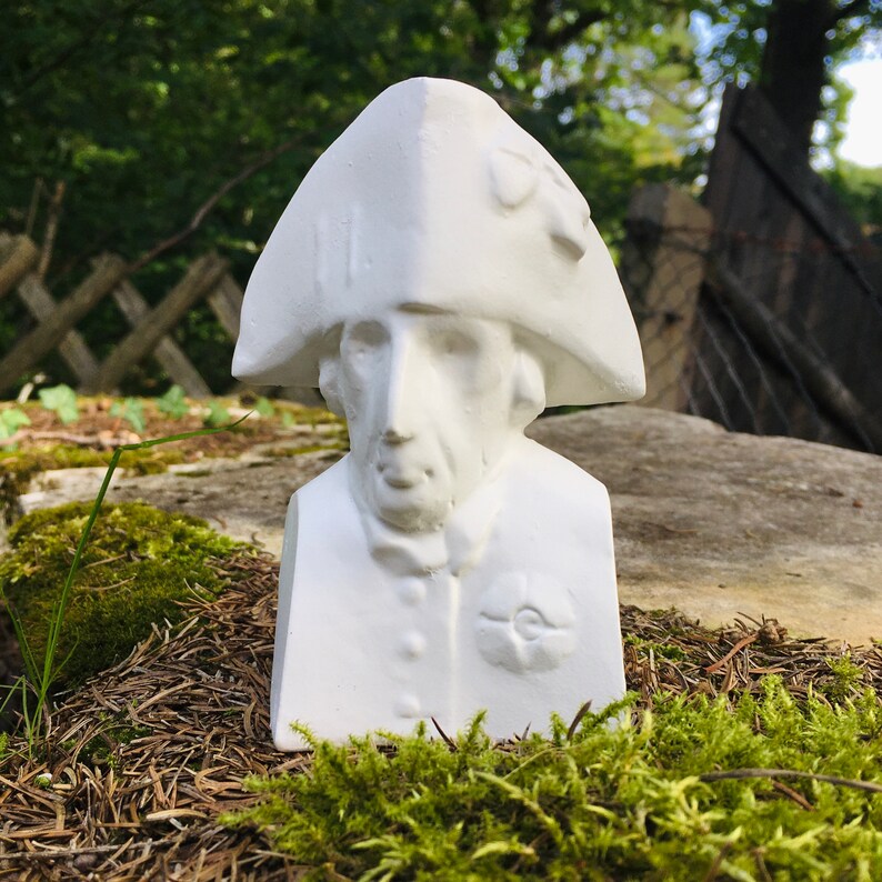 May include: White plaster bust of a person wearing a hat. The bust is sitting on a bed of moss and pine needles.