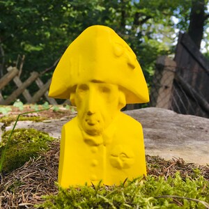 May include: A yellow bust sculpture of a person wearing a hat. The sculpture is made of a smooth, solid material and has a simple, classic design.