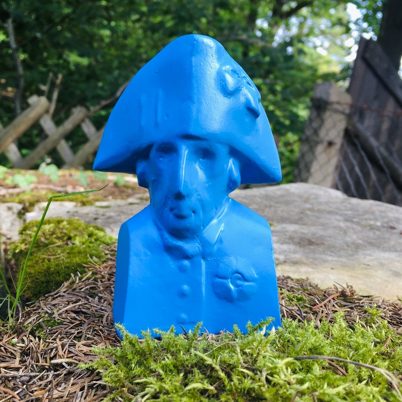 May include: A blue painted bust of a person wearing a blue hat. The bust is sitting on a bed of moss.
