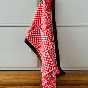 Puede incluir: Una toalla de cocina a cuadros rojos y blancos con un borde marrón y diseños florales blancos. La toalla está colgada de un gancho.