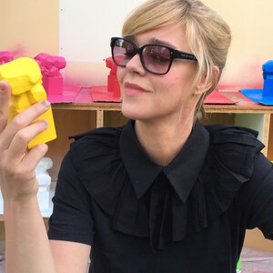 May include: A woman in sunglasses and a black shirt examines a bright yellow sculpture. The sculpture is a stylized bust. Other painted sculptures in pink, red, and blue are visible in the background.