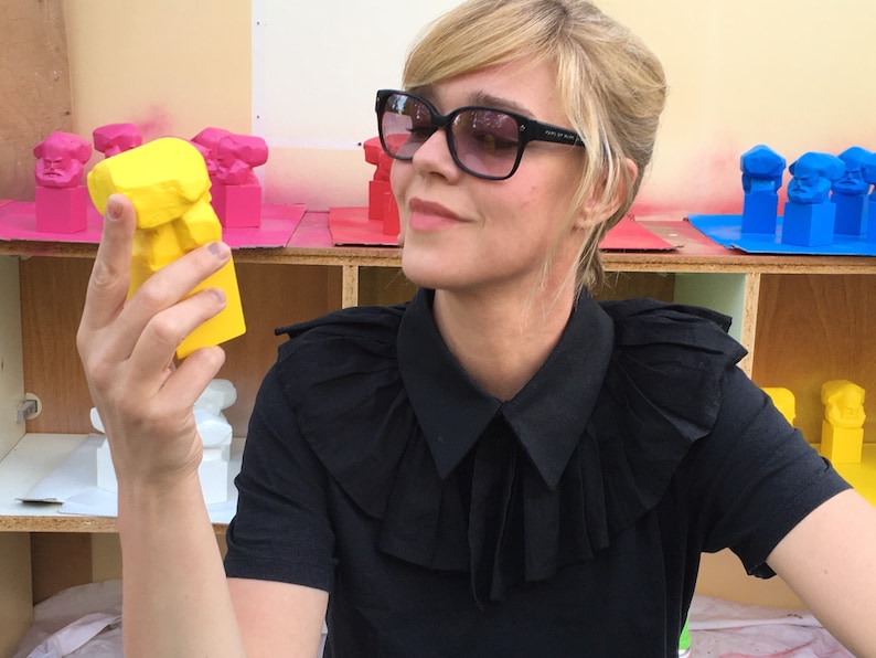 May include: A woman in sunglasses and a black shirt examines a bright yellow sculpture. The sculpture is a stylized bust. Other painted sculptures in pink, red, and blue are visible in the background.