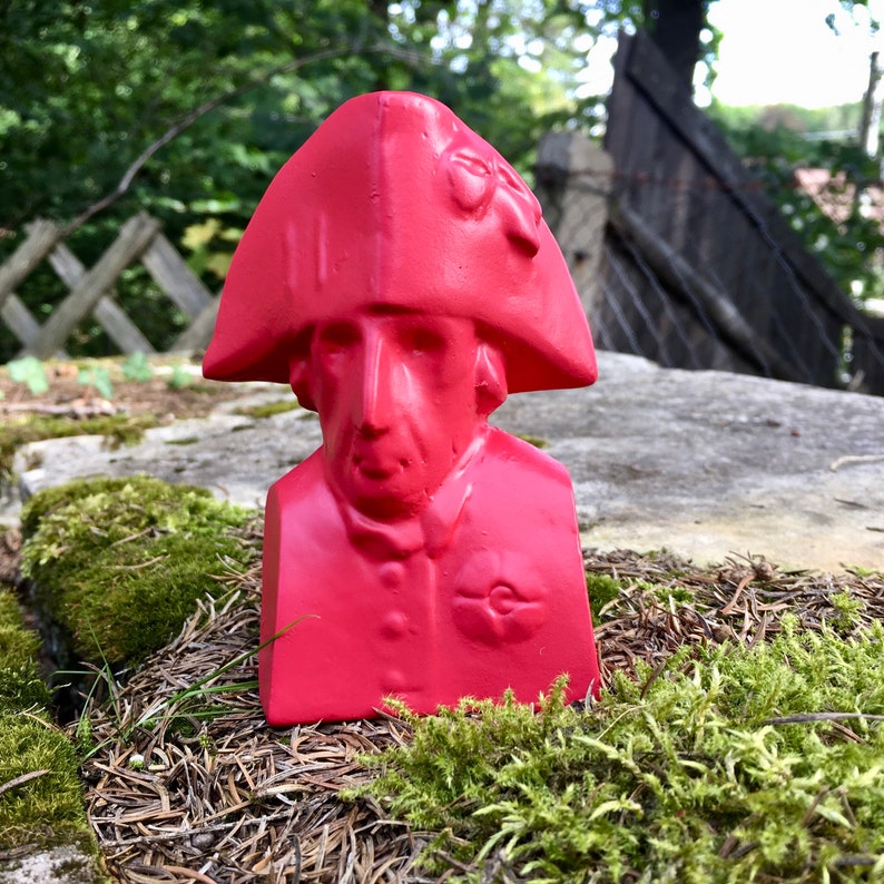 May include: A red bust of a person wearing a red hat with a decorative detail. The bust is sitting on a bed of moss.