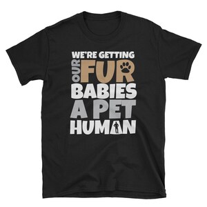 Puede incluir: Camiseta negra con el texto "We're getting our fur babies a pet human" en letras blancas y marrones. Una huella de pata est&aacute; incluida en el texto.