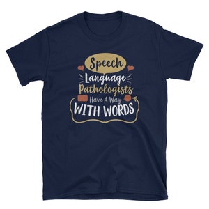 May include: Navy blue t-shirt with a white graphic that says "Speech Language Pathologists Have A Way With Words". The graphic includes a speech bubble, hearts, and a curved arrow.