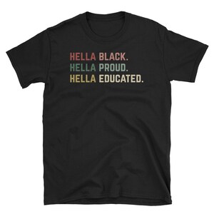 May include: Black t-shirt with the phrases "HELLA BLACK.", "HELLA PROUD.", and "HELLA EDUCATED." in a retro font. The text is in red, green, and gold colors. The shirt is made of a soft material.