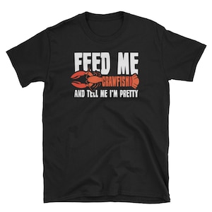 Peut inclure: T-shirt noir avec du texte blanc qui dit "Nourris-moi de crawfish et dis-moi que je suis belle". Une illustration de crawfish rouge est au-dessus du texte.