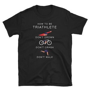 Op de afbeelding: Zwart T-shirt met witte tekst die "Hoe een triatleet te zijn" luidt en drie illustraties van een persoon die zwemt, fietst en rent met de tekst "Verdrink niet", "Crash niet" en "Loop niet" onder elke illustratie.