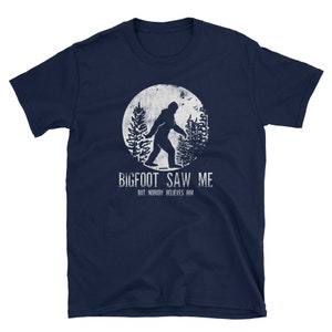 May include: A navy blue t-shirt with a white graphic of Bigfoot walking in a forest. The text "BIGFOOT SAW ME BUT NOBODY BELIEVES HIM" is printed below the graphic.