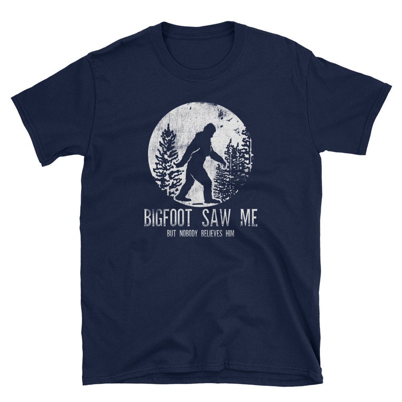 May include: A navy blue t-shirt with a white graphic of Bigfoot walking in a forest. The text "BIGFOOT SAW ME BUT NOBODY BELIEVES HIM" is printed below the graphic.