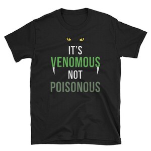 Op de afbeelding: Zwart T-shirt met een groene en witte afbeelding met de tekst "It's Venomous Not Poisonous" met twee gele ogen boven de tekst.