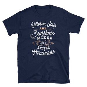 May include: Navy blue t-shirt with the text "October Girls are Sunshine Mixed with a Little Hurricane" in white and peach-colored script. The shirt has a crew neck and short sleeves.