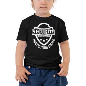 以下が含まれることがあります： 白の盾のグラフィックが描かれた黒い幼児用Tシャツ。"Security Big Brother Protection Squad"と書いてあります。