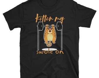 Op de afbeelding: Zwart T-shirt met een cartoon oranje kat die gewichten heft. De tekst "Kitten my swole on" is op het T-shirt gedrukt.