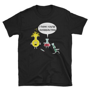 Puede incluir: Camiseta negra con un diseño de dibujos animados de cristalería de laboratorio. La imagen muestra un matraz amarillo, un vaso de precipitados rojo y un tubo de ensayo verde, con el texto "I THINK YOU'RE OVERREACTING" en una burbuja. El diseño es humorístico.