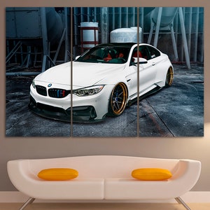 K&ouml;nnte beinhalten: Ein wei&szlig;er BMW M4 mit goldenen Felgen, der in einer industriellen Umgebung geparkt ist. Das Auto hat einen schwarzen Frontsplitter und Seitenschweller. Das Auto ist scharf, w&auml;hrend der Hintergrund unscharf ist.