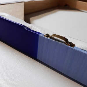 K&ouml;nnte beinhalten: Nahaufnahme eines blauen und wei&szlig;en Leinwandgem&auml;ldes mit einem goldenen Metall-Aufh&auml;nger an der R&uuml;ckseite.