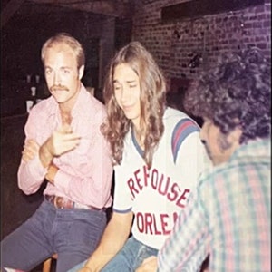 May include: Three people are standing together. The person in the middle is wearing a white shirt with red and blue trim and the text "REFUSE ORLEANS" on the front. The person on the left is wearing a pink button-down shirt and blue jeans. The person on the right is wearing a plaid shirt.