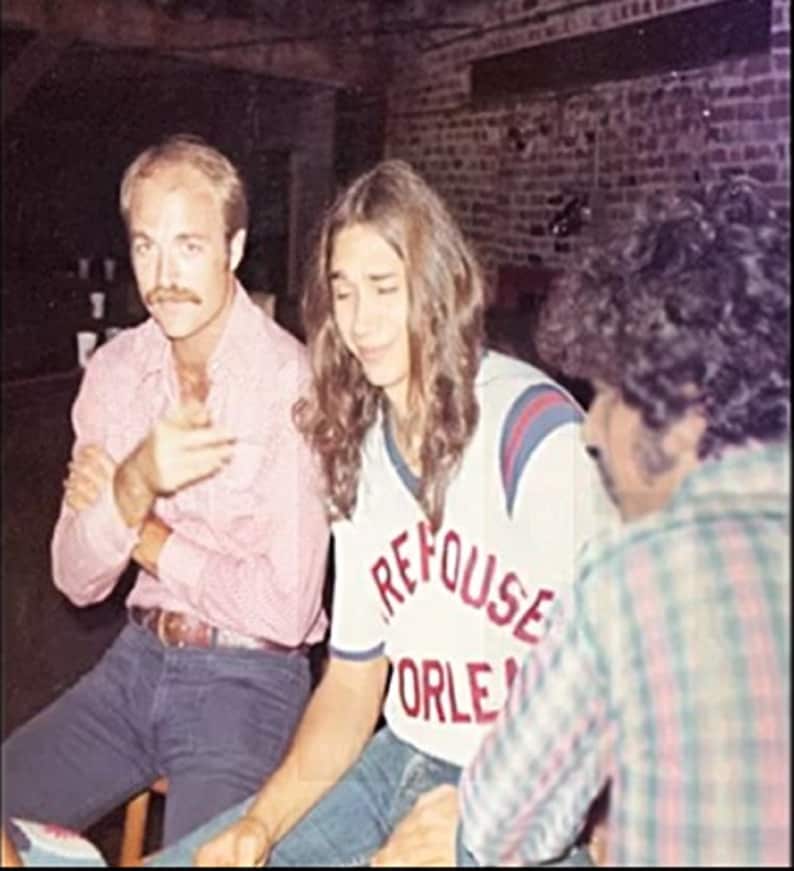 May include: Three people are standing together. The person in the middle is wearing a white shirt with red and blue trim and the text "REFUSE ORLEANS" on the front. The person on the left is wearing a pink button-down shirt and blue jeans. The person on the right is wearing a plaid shirt.