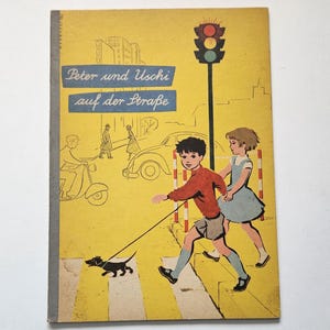 Pode incluir: Uma capa de livro amarela com uma ilustração de duas crianças a passear um cão numa passadeira. O título do livro é "Peter und Uschi auf der Strabe".