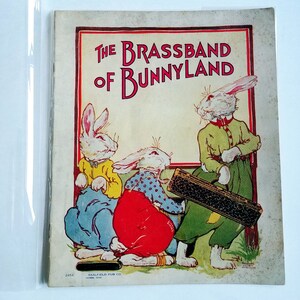Pode incluir: Uma capa de livro infantil vintage com três coelhos brancos de orelhas compridas. Os coelhos estão vestidos com roupas coloridas e segurando um instrumento musical. O título do livro é "The Brassband of Bunnyland".