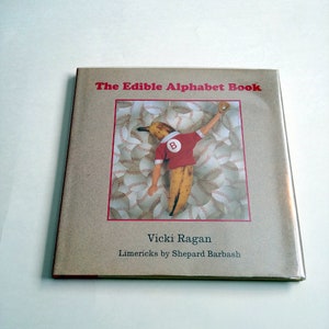 Puede incluir: Un libro infantil titulado "The Edible Alphabet Book" con una ilustración de portada de una figura de plátano con uniforme de béisbol. El fondo presenta pelotas de béisbol. La autora es Vicki Ragan, con limericks de Shepard Barbash.