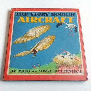 May include: The Story Book of Aircraft by Maud and Miska Petersham. The cover features a blue sky with a white stork flying and a yellow glider with a man flying in it. There is a nest with a white stork in it on the bottom right corner.
