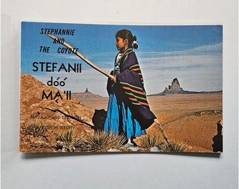 May include: A vintage postcard featuring a young person in traditional Native American clothing, holding a long stick, set against a desert landscape. The postcard's text reads "Stephannie and the Coyote" and "Stefanii dóó Ma'ii" in Navajo and English.