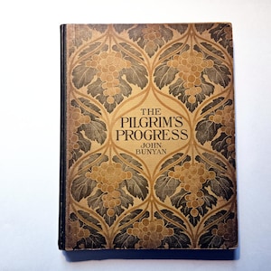 May include: A vintage hardcover book titled "The Pilgrim's Progress" by John Bunyan. The cover features a detailed Art Nouveau design with grape clusters and leaves in shades of brown and gold against a tan background. The book's spine is black.